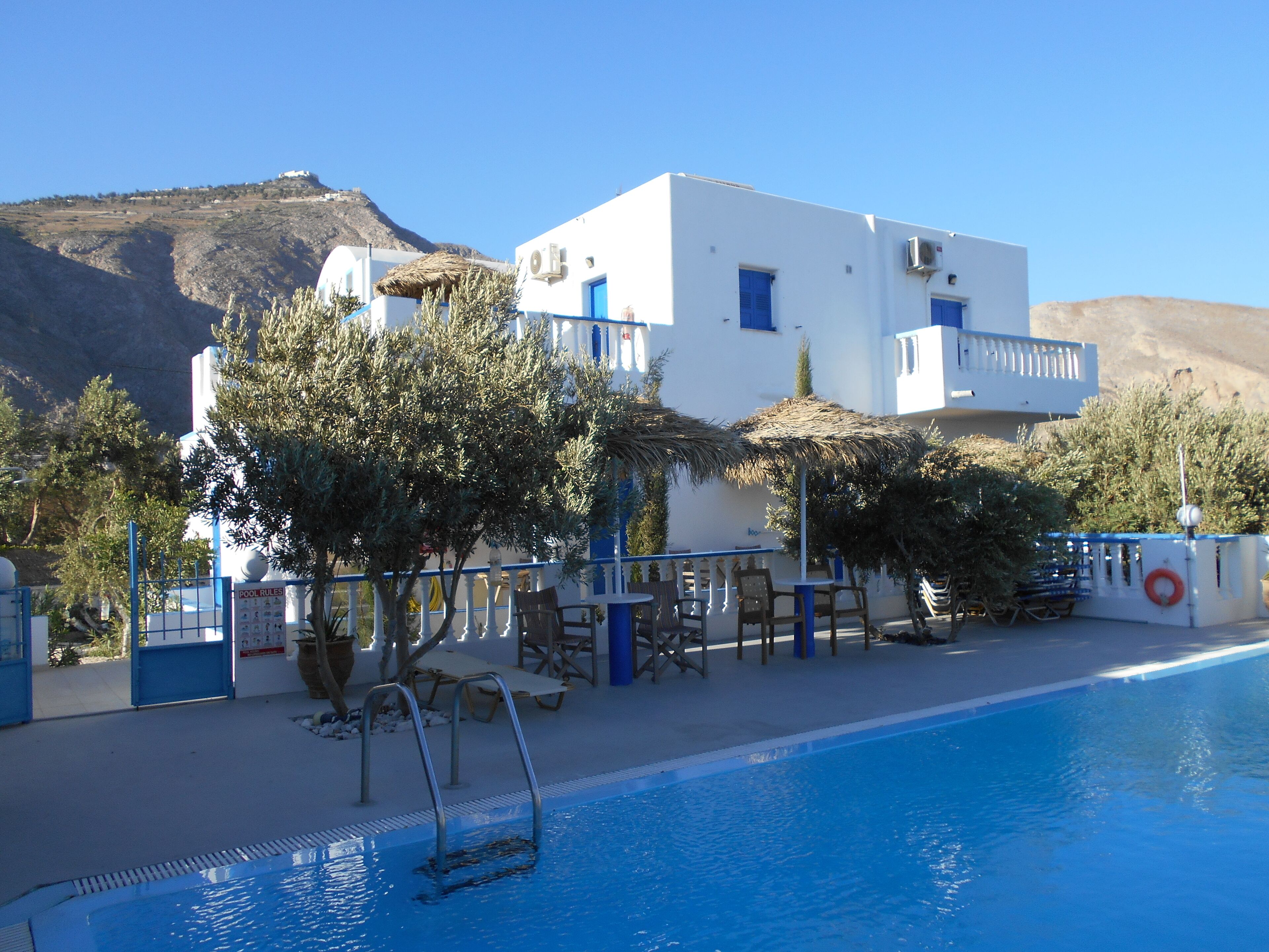 Zona de piscina con una piscina azul cristalina, edificios blancos con detalles azules y un paisaje montañoso de fondo.