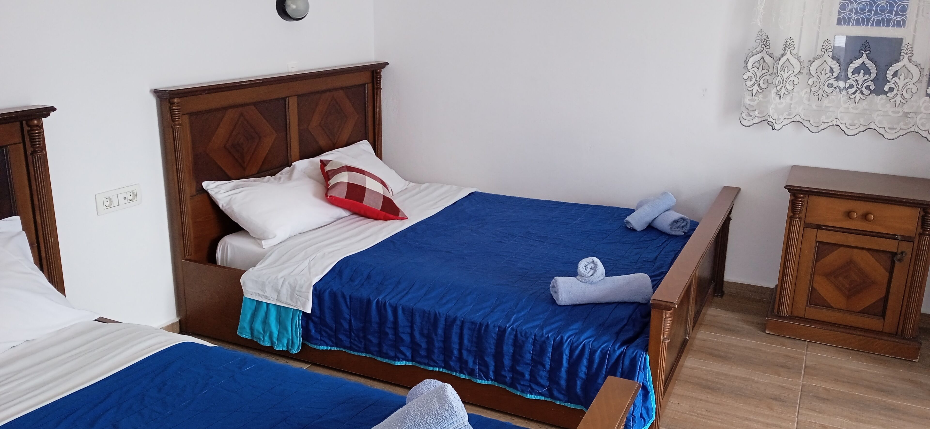 A hotel room with two beds, each with a blue bedspread and white pillows. There is a wooden nightstand with a drawer and a decorative mirror on the wall.
