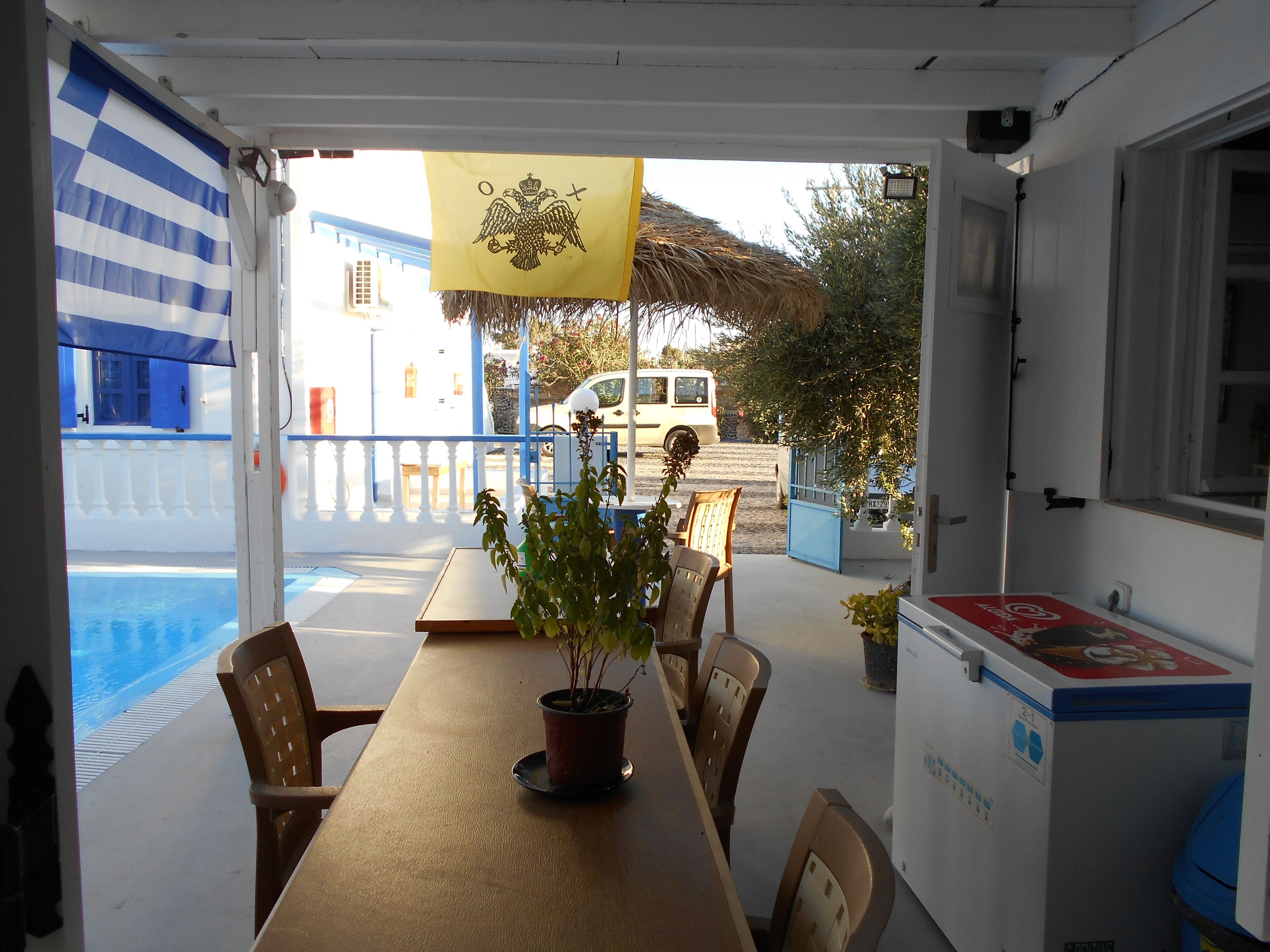 A covered outdoor seating area with a table, chairs, and a potted plant. A white van is parked outside, and a building with blue and white walls is visible in the background.