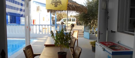 A covered outdoor seating area with a table, chairs, and a potted plant. A white van is parked outside, and a building with blue and white walls is visible in the background.