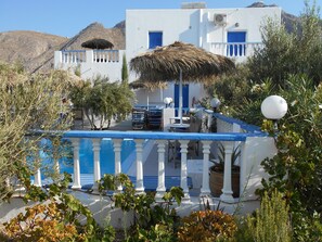 A white building with blue accents, a pool, and a thatched roof. - Studios Apartments Kapetanios (Santorini)