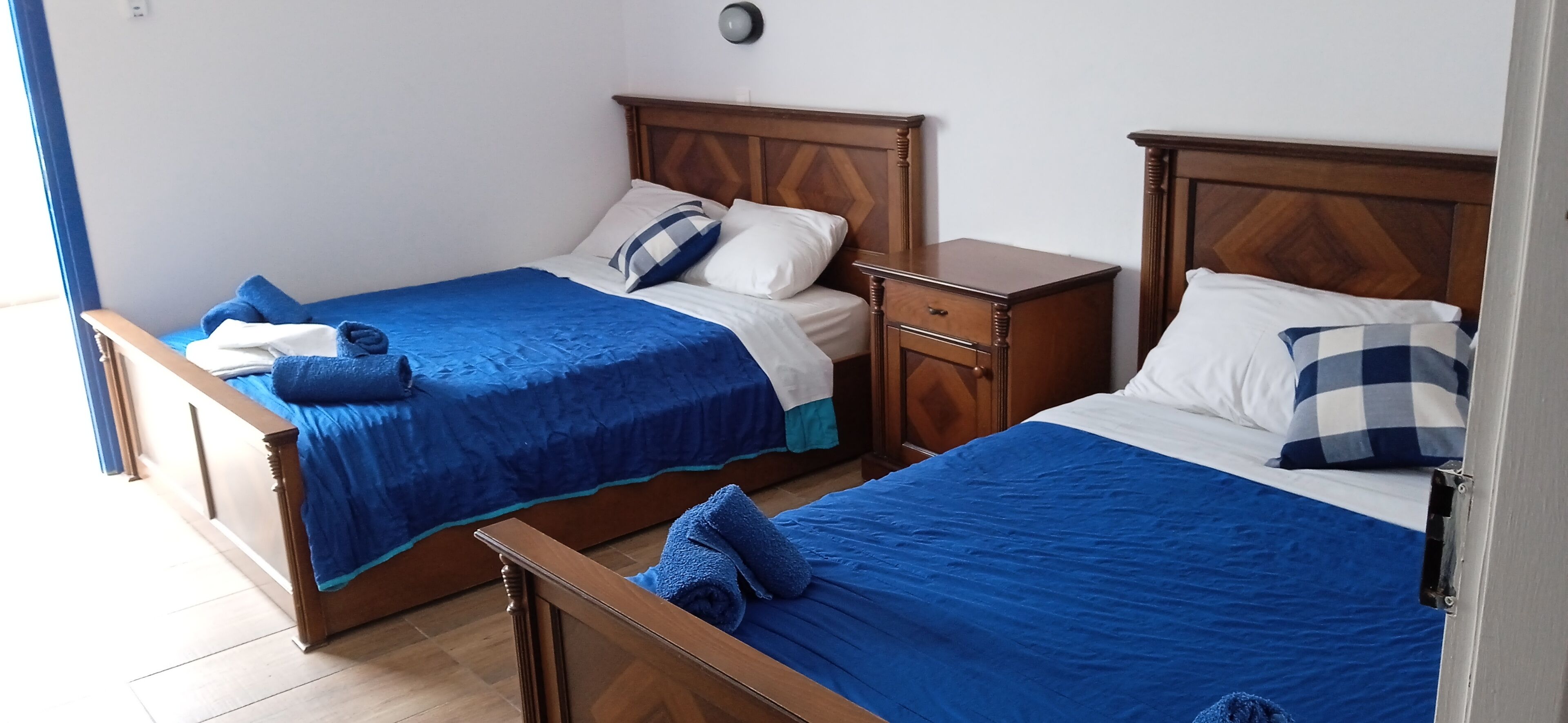 Twee houten bedden met blauw beddengoed, een houten nachtkastje en een wandlamp.
