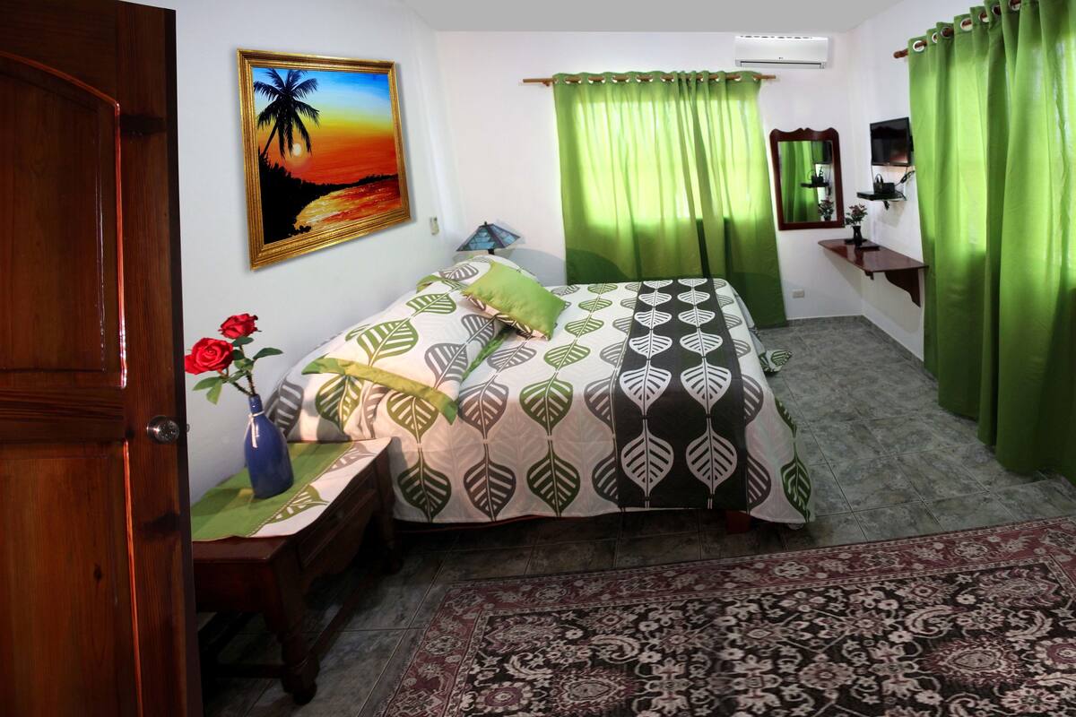 A bedroom with a bed, a nightstand, a vase with flowers, a painting, a mirror, and green curtains.