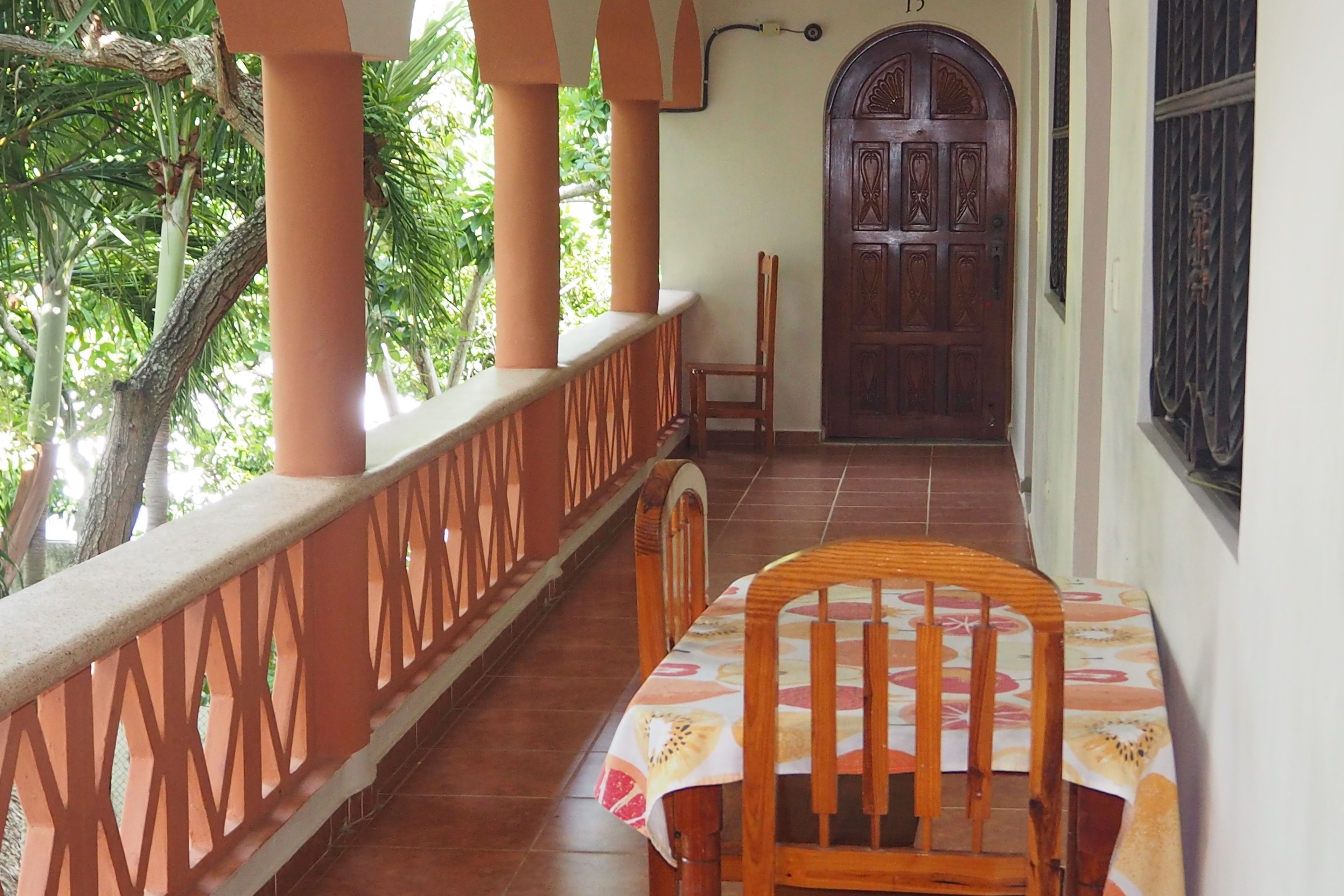 A covered outdoor area with a wooden table and chairs, a door, and a view of greenery.