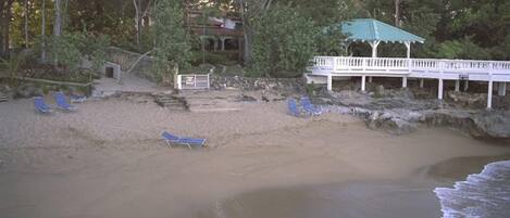 A beach with a white gazebo, blue lounge chairs, and a building with a green roof.