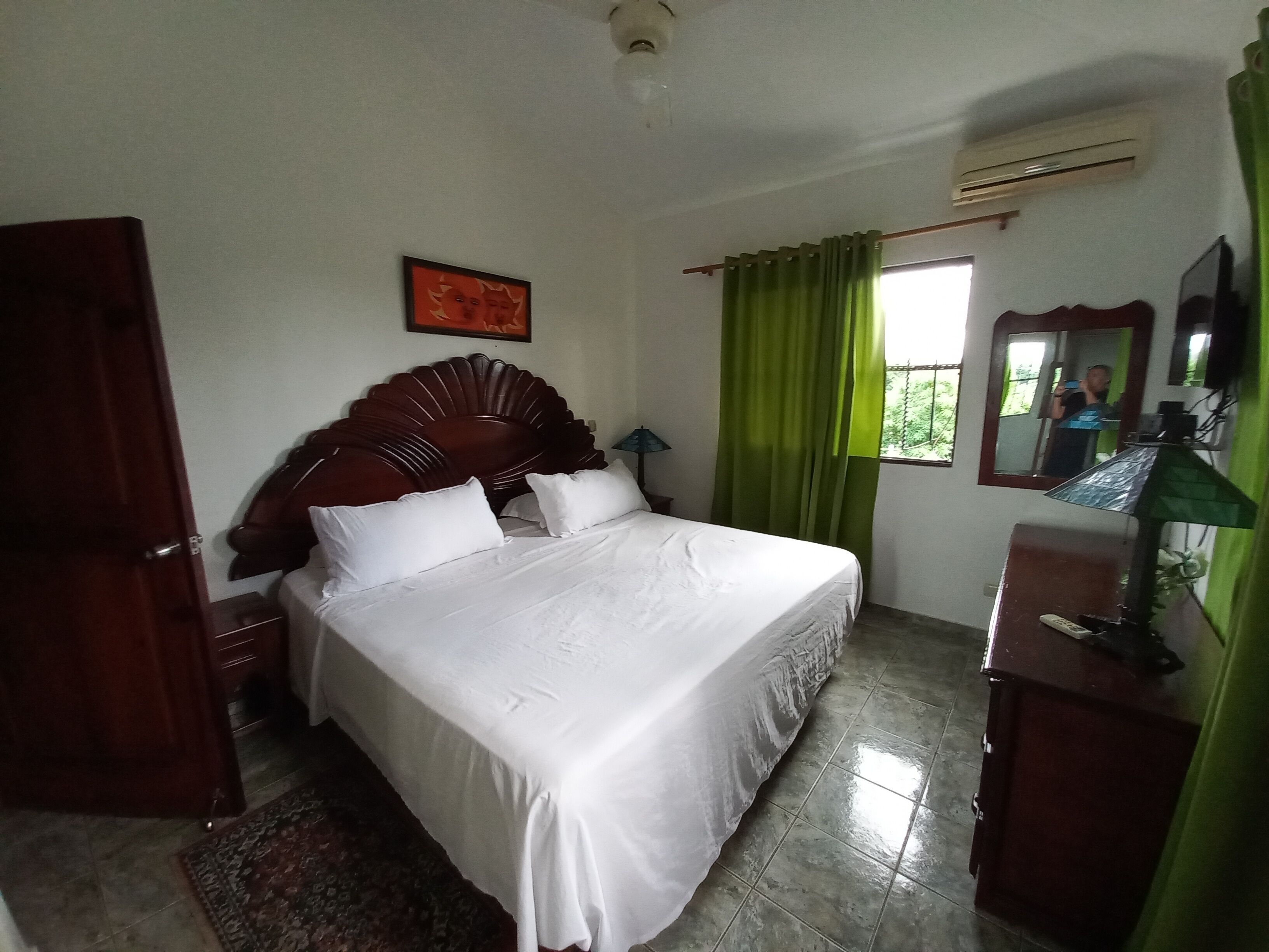 Cama bem arrumada com lençóis brancos, cabeceira de madeira, mesa de cabeceira com abajur, cortina verde, espelho e pequena escrivaninha com abajur.
