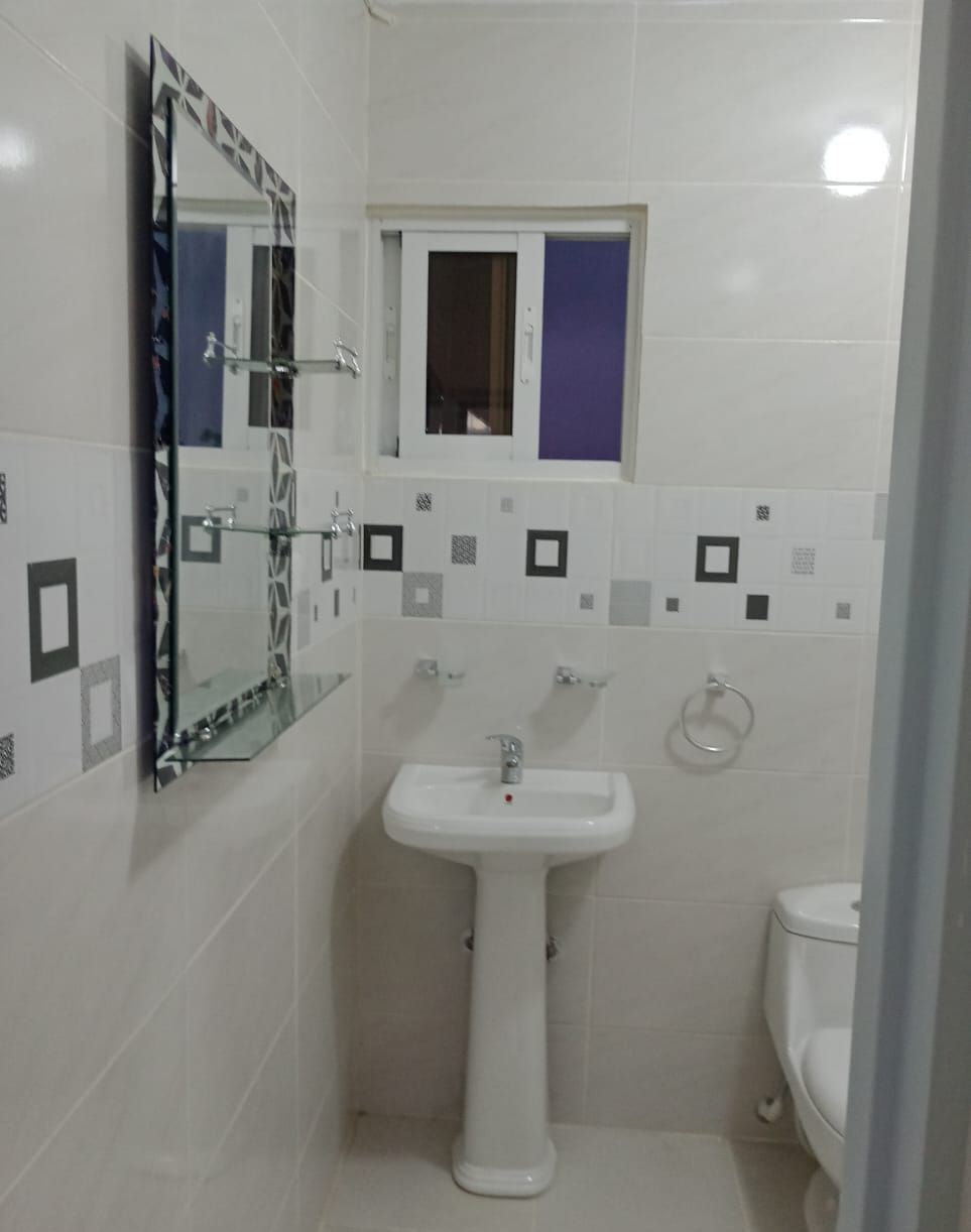 Een compacte, schone badkamer met een witte wastafel, een spiegel met geometrisch vormgegeven lijst en een raam boven de wastafel.