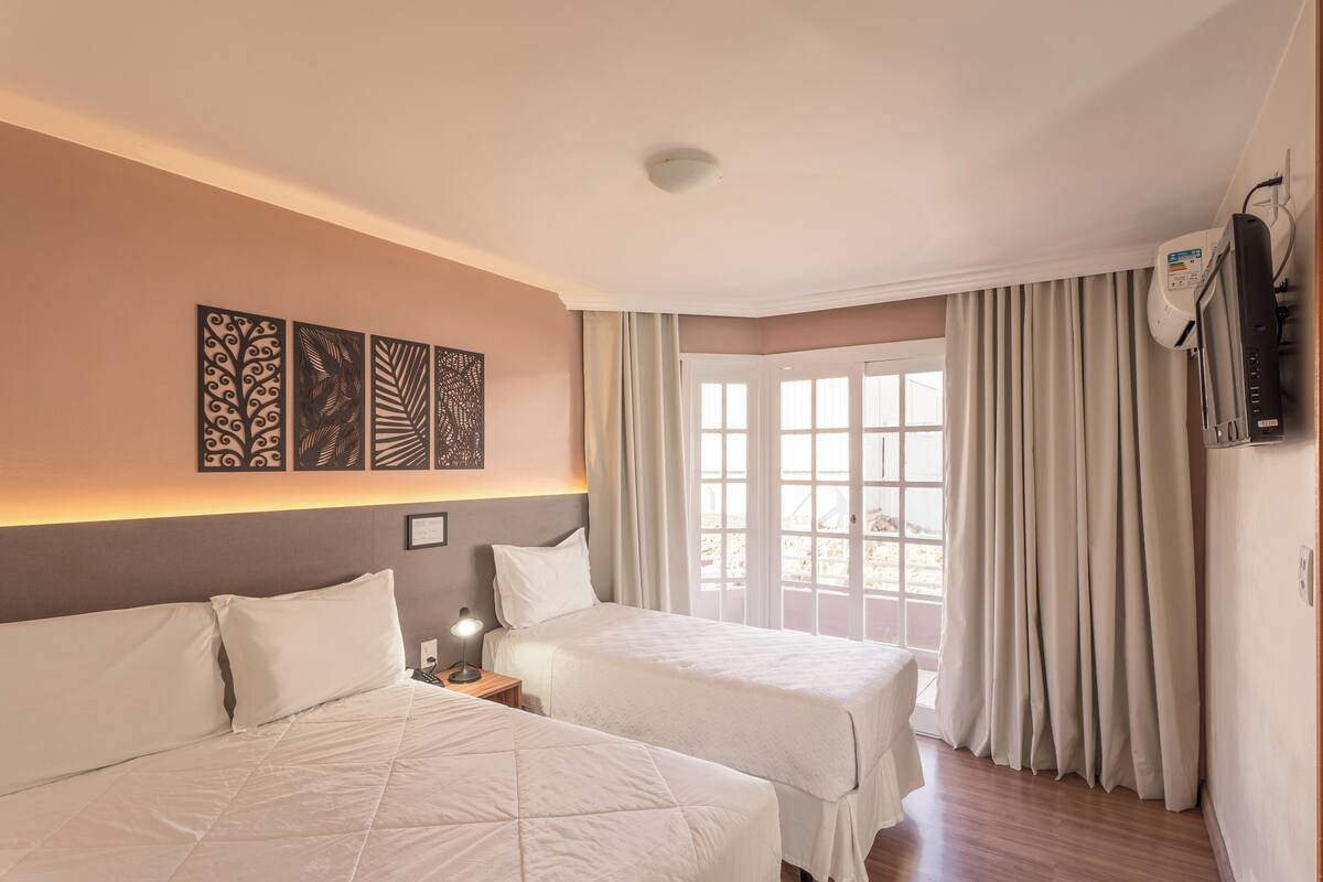 Une chambre d’hôtel avec deux lits, un luminaire au plafond, un téléviseur fixé au mur et une fenêtre avec des rideaux.