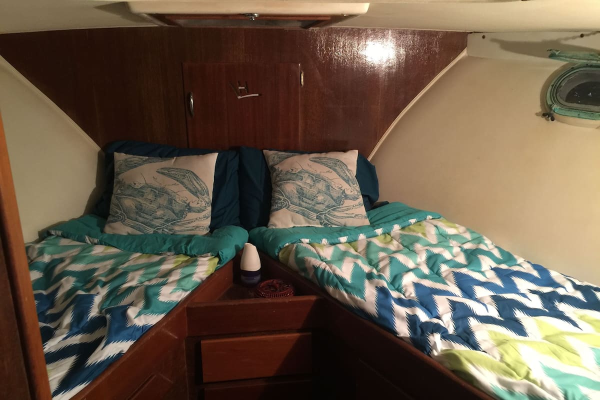 A small, compact bedroom with two single beds, each with patterned bedding. There is a small wooden dresser and a small table with a bottle on it.