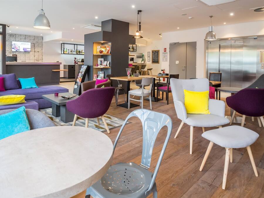 Eine moderne Hotelhalle mit einer Rezeption, einer Sitzecke mit lilafarbenen und grauen Sofas sowie einem Essbereich mit Holztisch und Stühlen.