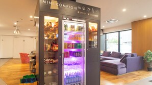 A mini kitchenette with a refrigerator stocked with snacks and drinks, a seating area with a purple sofa, and a wooden floor. - Campanile PRIME - Metz Centre Gare (Metz)