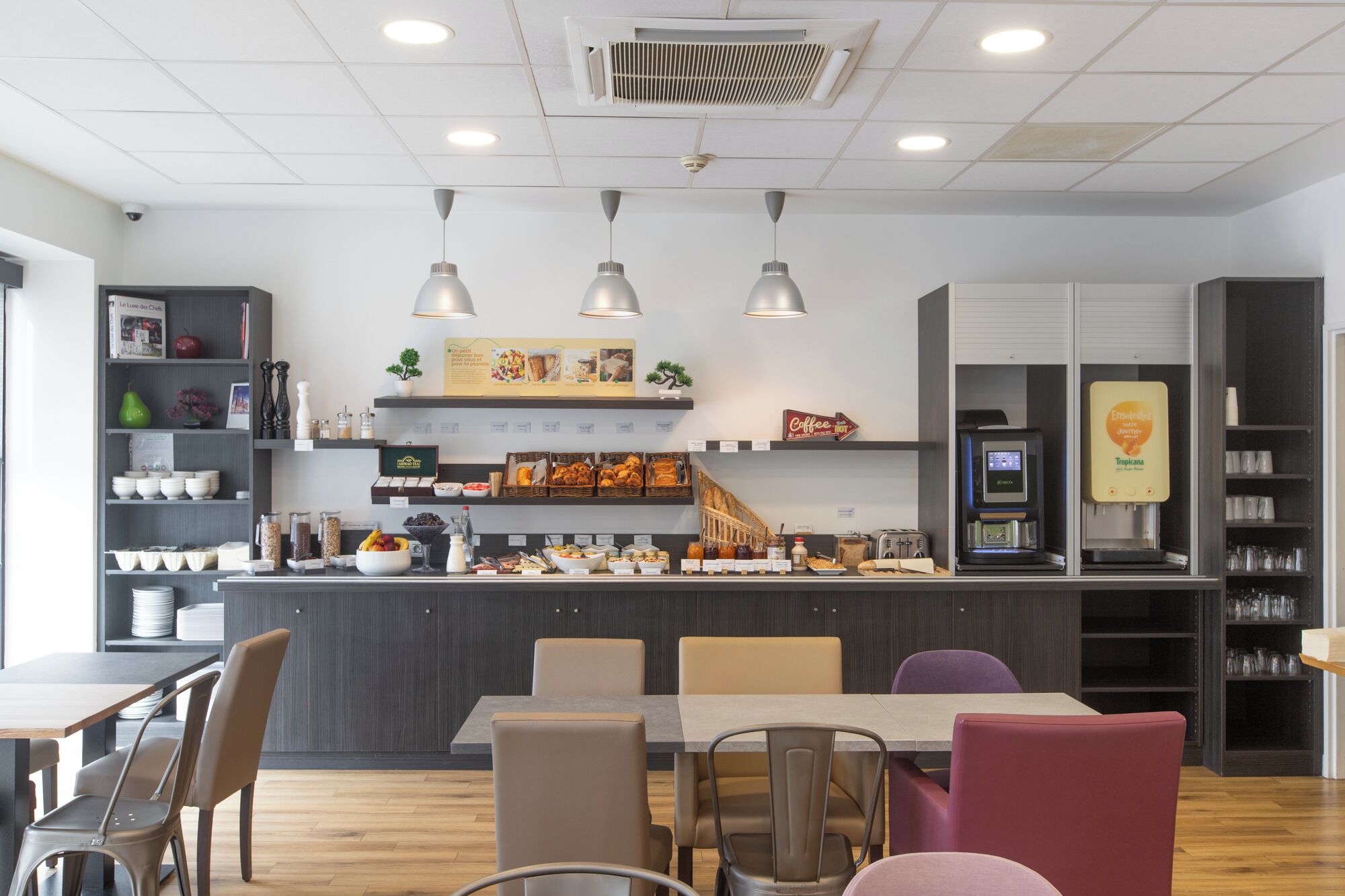 A modern cafe with a counter displaying various food items and a coffee machine. There are multiple seating options including tables and chairs.