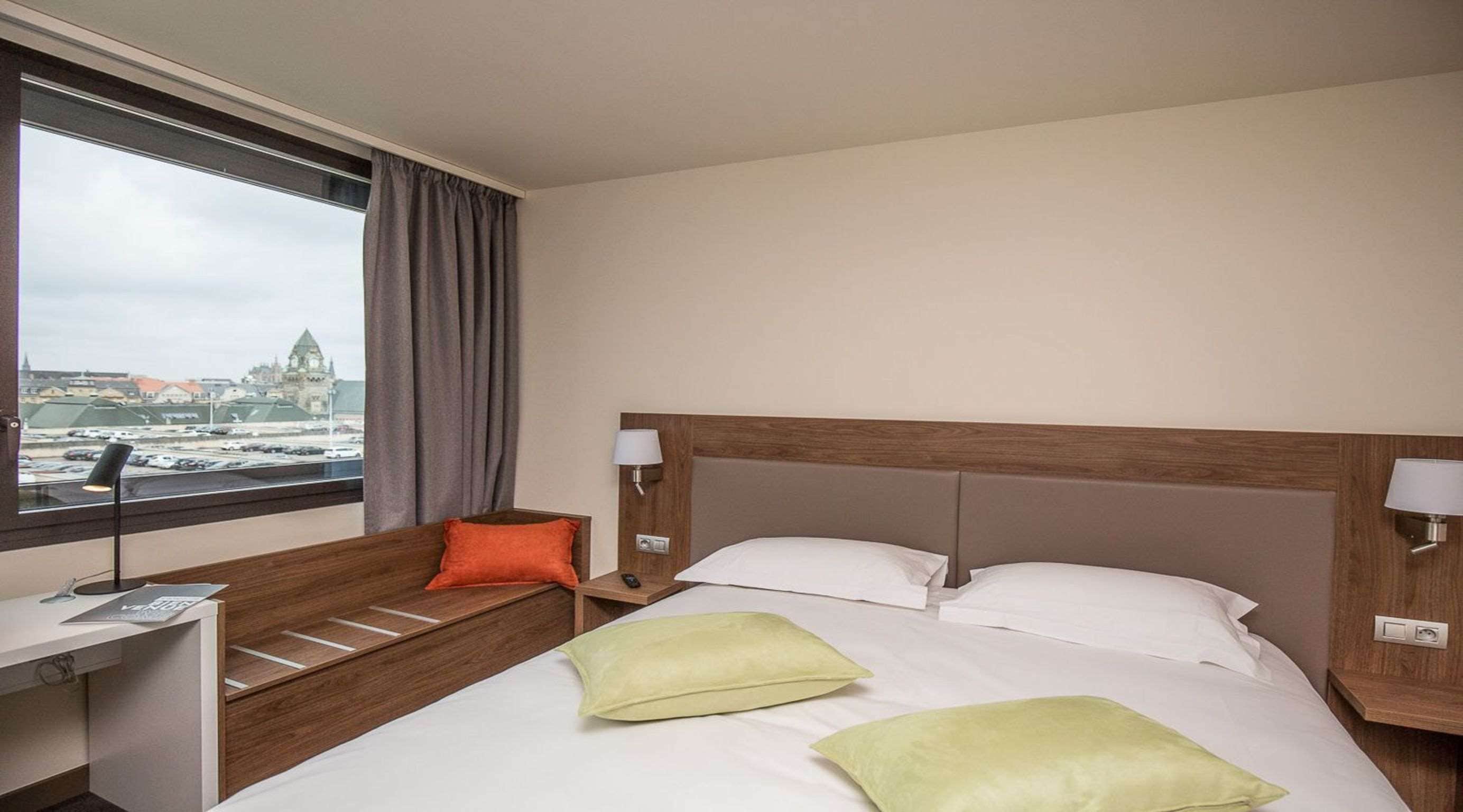 Ein Hotelzimmer mit Bett, Schreibtisch, Stuhl, Fenster mit Blick auf eine Stadtlandschaft und einer Lampe.