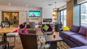 A modern hotel lobby with a television, seating area, and large windows offering a city view. - Campanile PRIME - Metz Centre Gare (Metz)