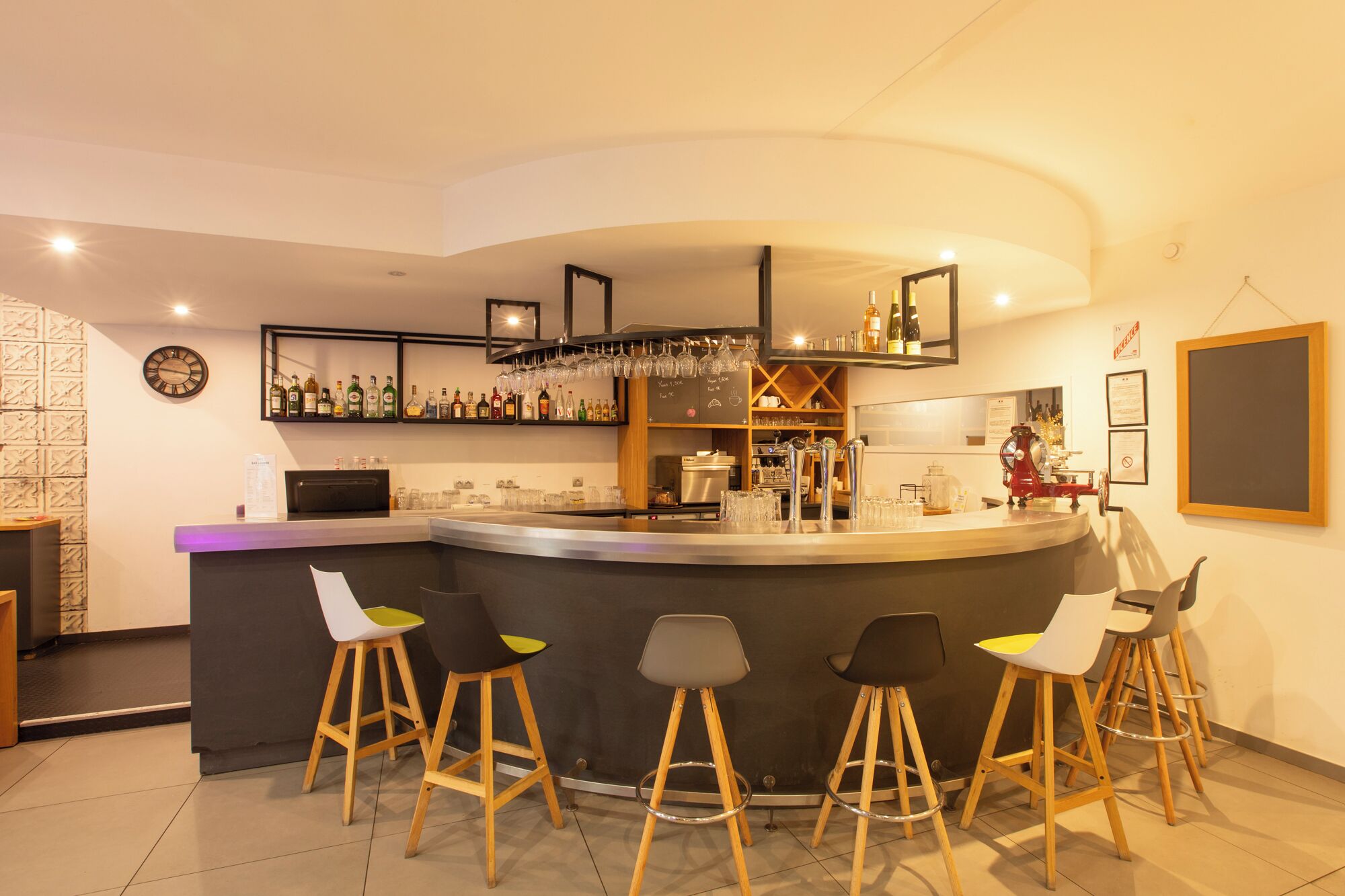 Zona de bar moderna con mostrador curvo, taburetes, estantes con botellas de licor y una pizarra con la carta.