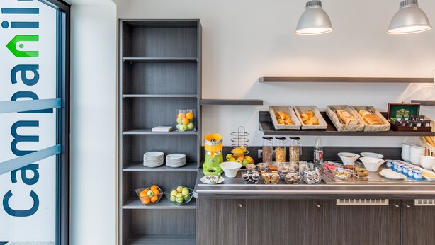 A breakfast buffet area with shelves of fruits, a juice dispenser, and various food items. - Campanile PRIME - Metz Centre Gare (Metz)