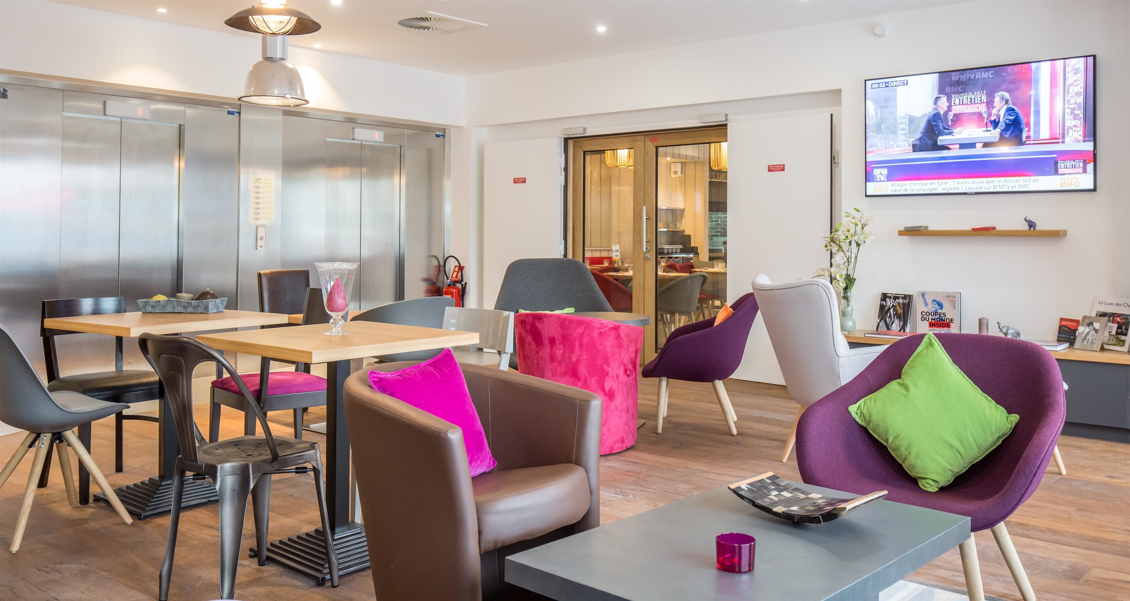 A modern hotel lobby with a dining area, a TV screen, and an elevator.