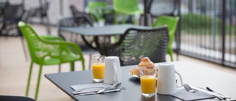 Una mesa dispuesta para el desayuno con jugo, un croissant y una taza de café, rodeada de sillas verdes y una cerca de metal.