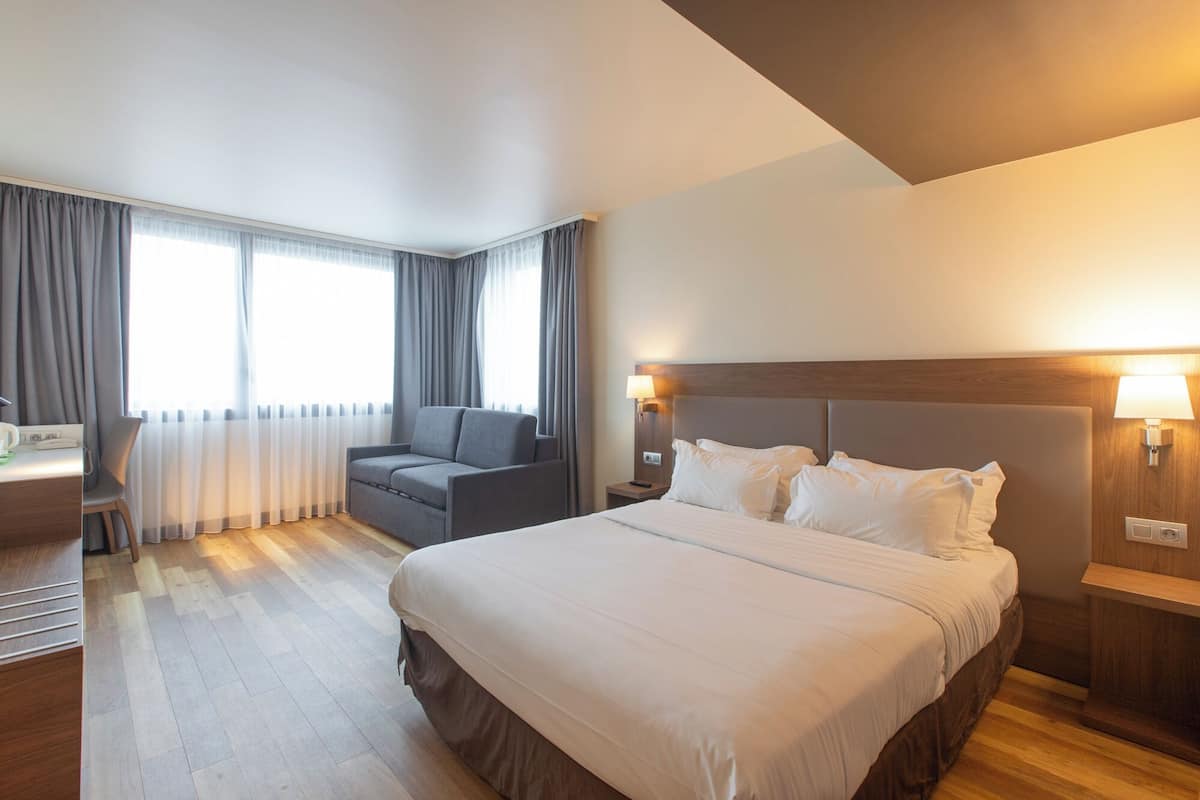 Habitación de hotel con una cama grande, un sofá, un escritorio con lámpara y una ventana con cortinas.
