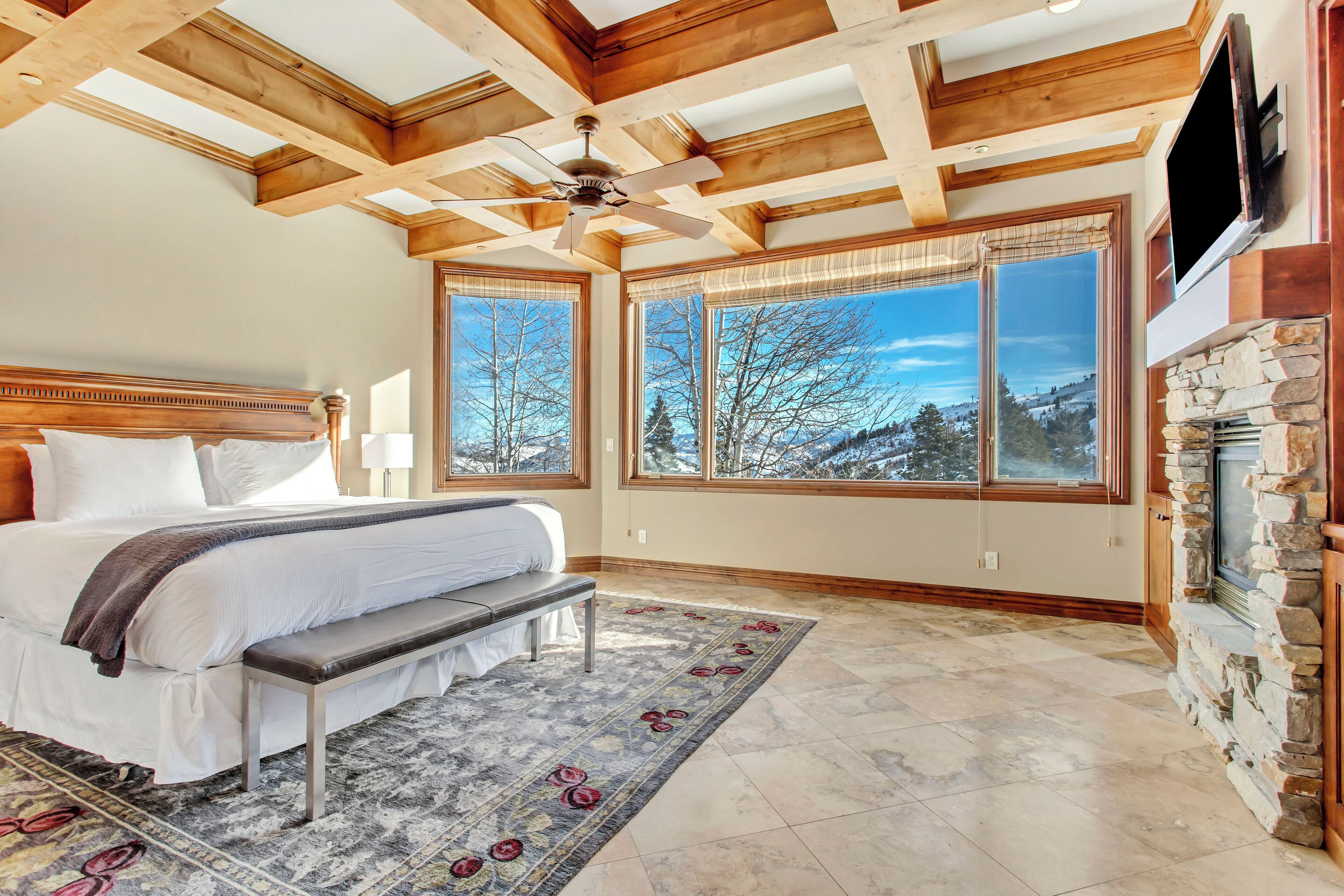 Un dormitorio con una cama grande, un banco, una chimenea y ventanas amplias con vistas a árboles y montañas.