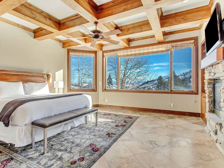 Un dormitorio con una cama grande, un banco, una chimenea y ventanas amplias con vistas a árboles y montañas.