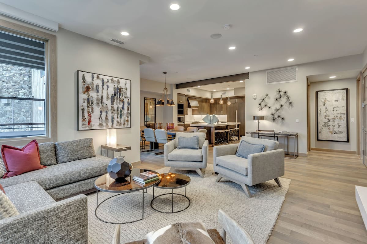 Un salon moderne avec un canapé gris, des fauteuils et une table basse. On y trouve également un coin repas avec une table et des chaises, et une cuisine à l’arrière-plan.