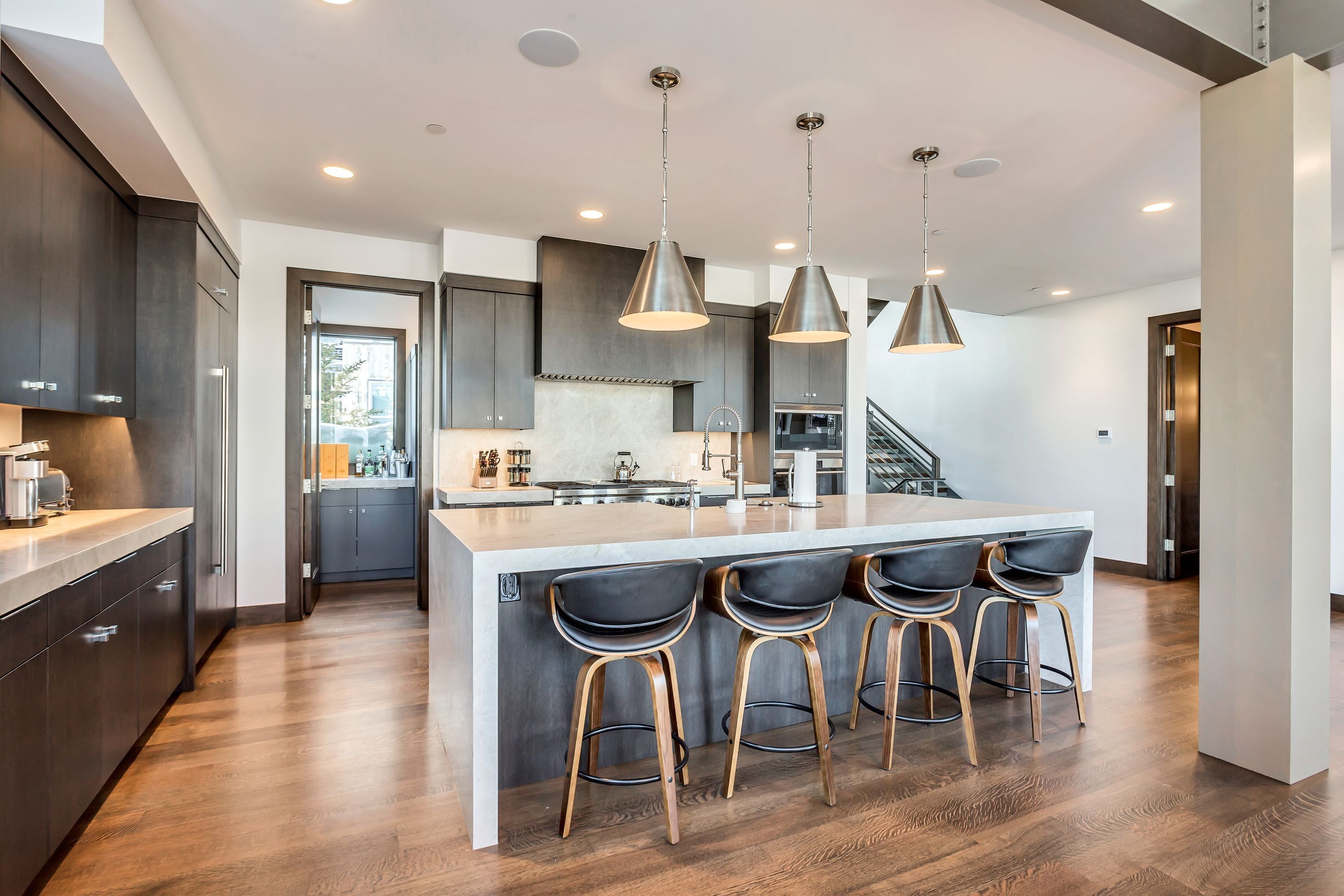 A modern kitchen with dark cabinetry, a central island with bar stools, pendant lighting, and a visible staircase.