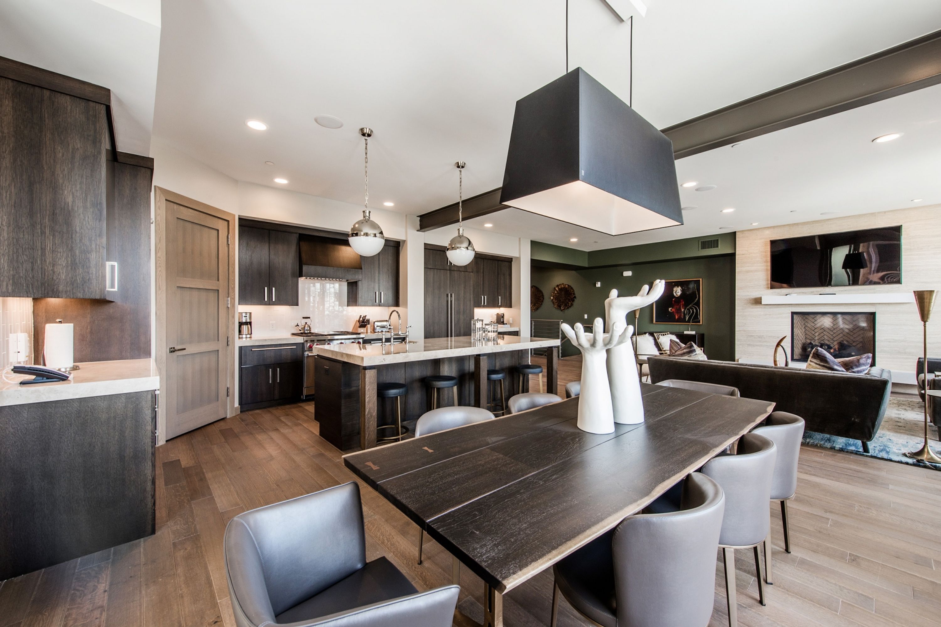 A modern kitchen with dark wood cabinets, a central island with bar stools, pendant lighting, and a dining area with a dark wood table and grey chairs.