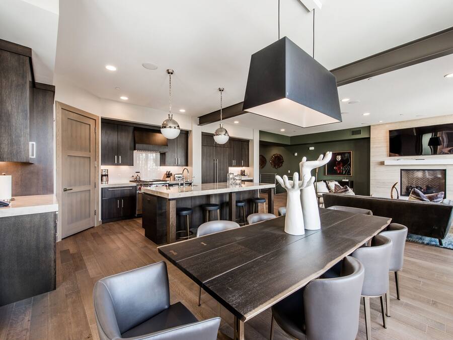 Una cocina moderna con armarios de madera oscura, una isla central con taburetes de bar, iluminación colgante y un comedor con mesa de madera oscura y sillas grises.