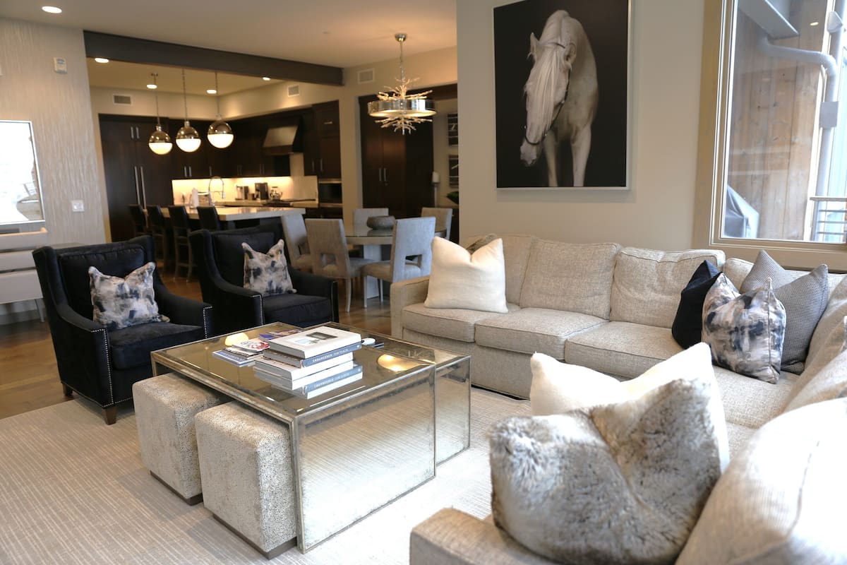 Un salon moderne avec un canapé, des fauteuils et des poufs. On y trouve une table basse en verre, sur laquelle sont posés des livres, et un lustre au-dessus.