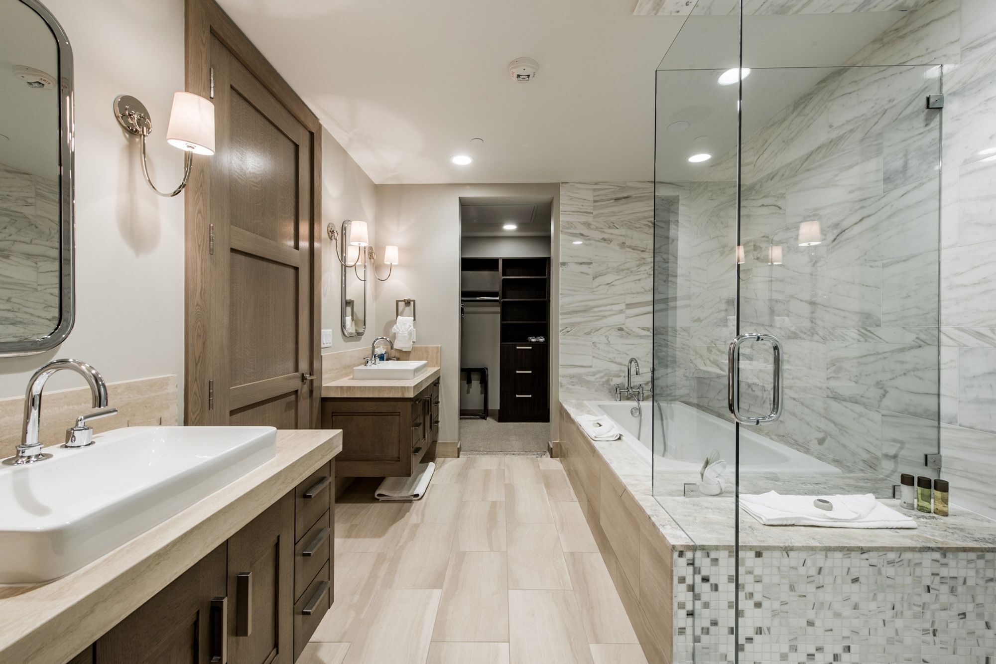 A modern bathroom with a large mirror, two sinks, a walk-in shower, and a walk-in closet.