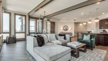 A modern living room with a sectional sofa, a coffee table, and a green armchair. There is a kitchen area with a dining table and pendant lighting.