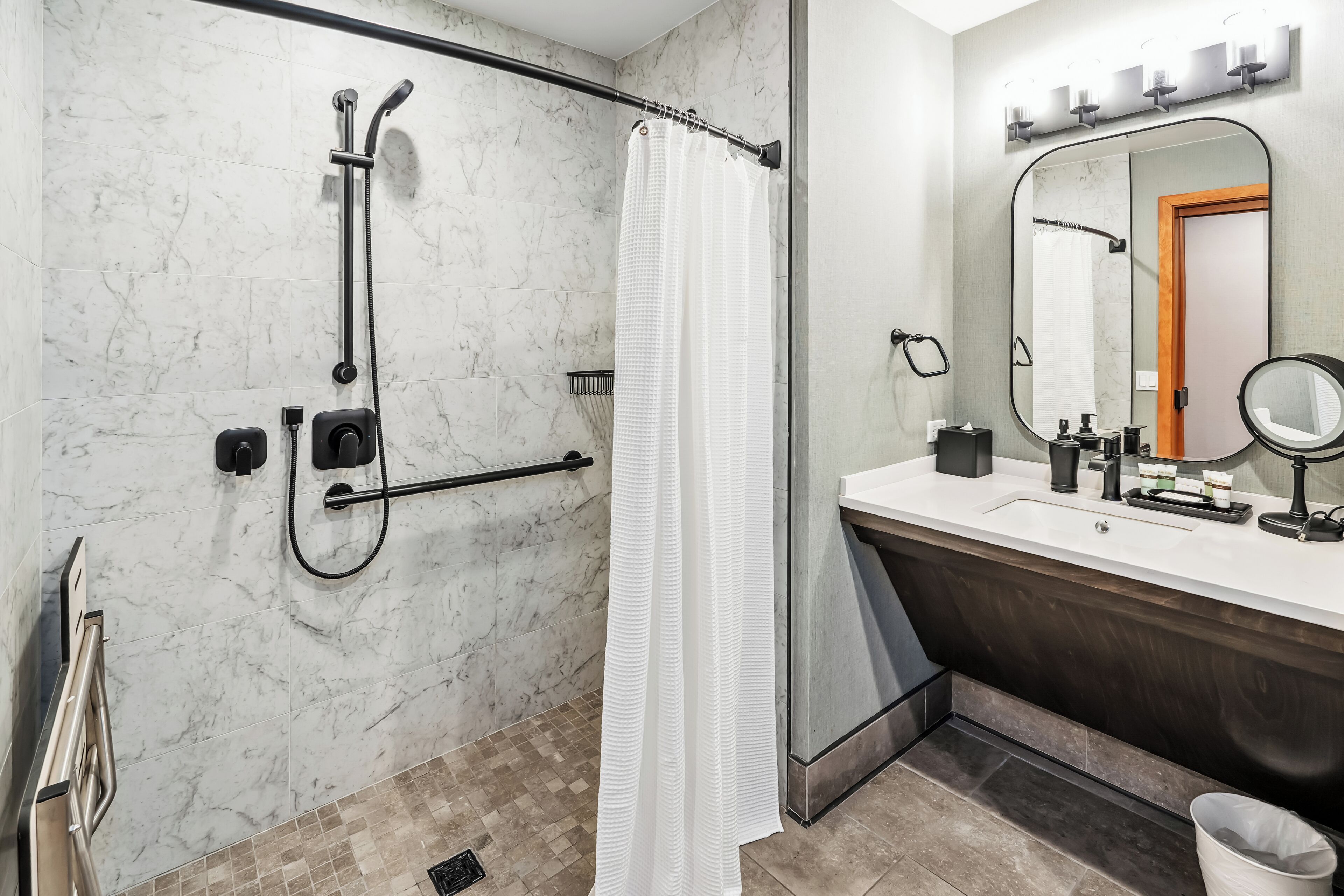 A modern bathroom with a walk-in shower, a white shower curtain, a large mirror, and a vanity with toiletries.