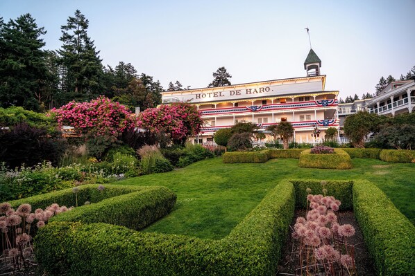 A hotel with a classic architectural style, featuring a prominent sign and a well-manicured garden with hedges and flowering plants.