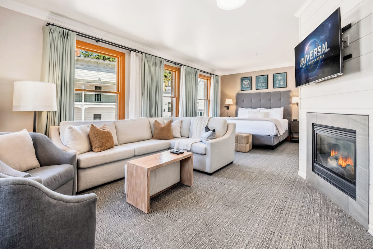 A modern hotel room with a sofa, armchair, and a bed. There is a fireplace and a television mounted on the wall.