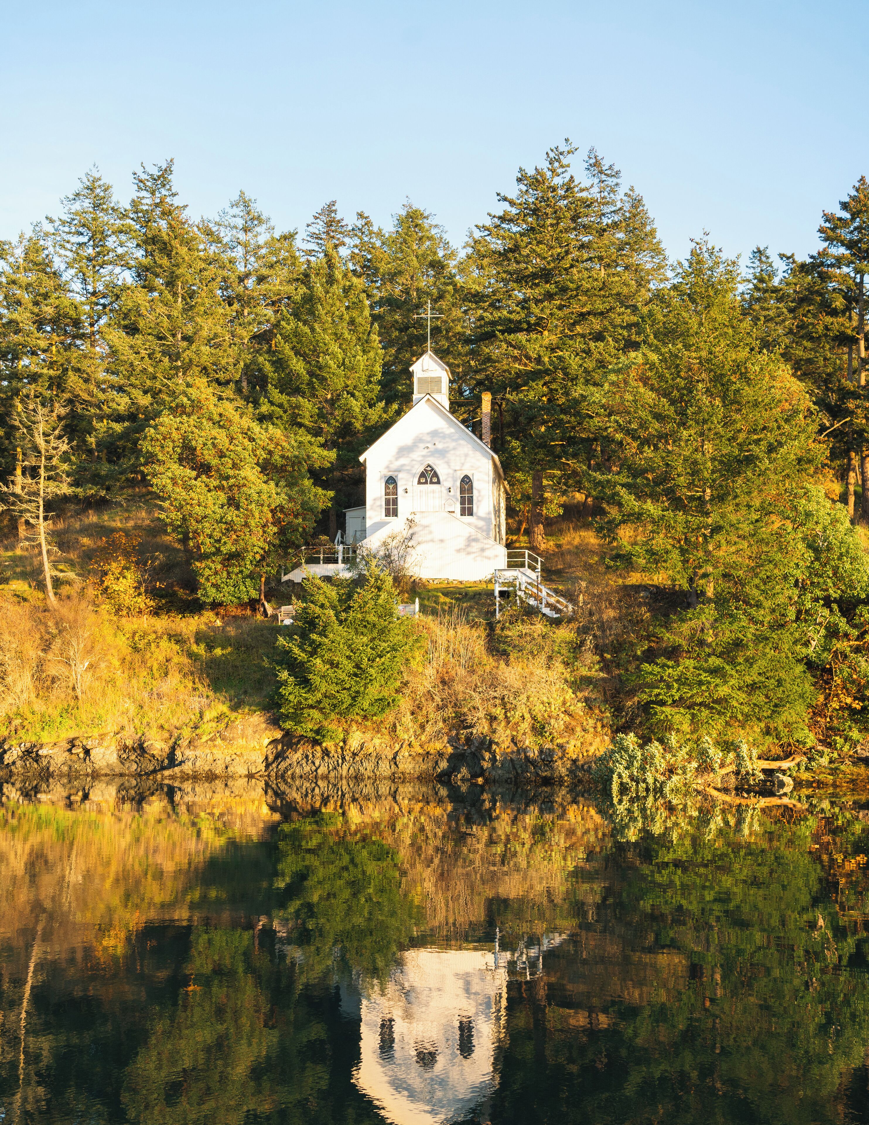 Uma igreja branca com um campanário, rodeada por árvores densas e refletida em águas calmas.