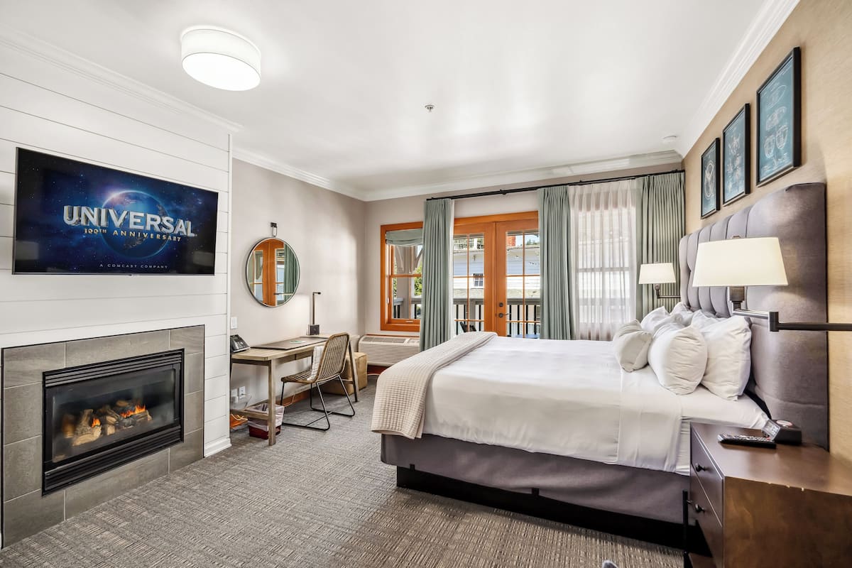 A hotel room with a large flat-screen TV displaying the Universal 100th Anniversary logo, a fireplace, a bed with a headboard, a desk with a chair, and a window with curtains.