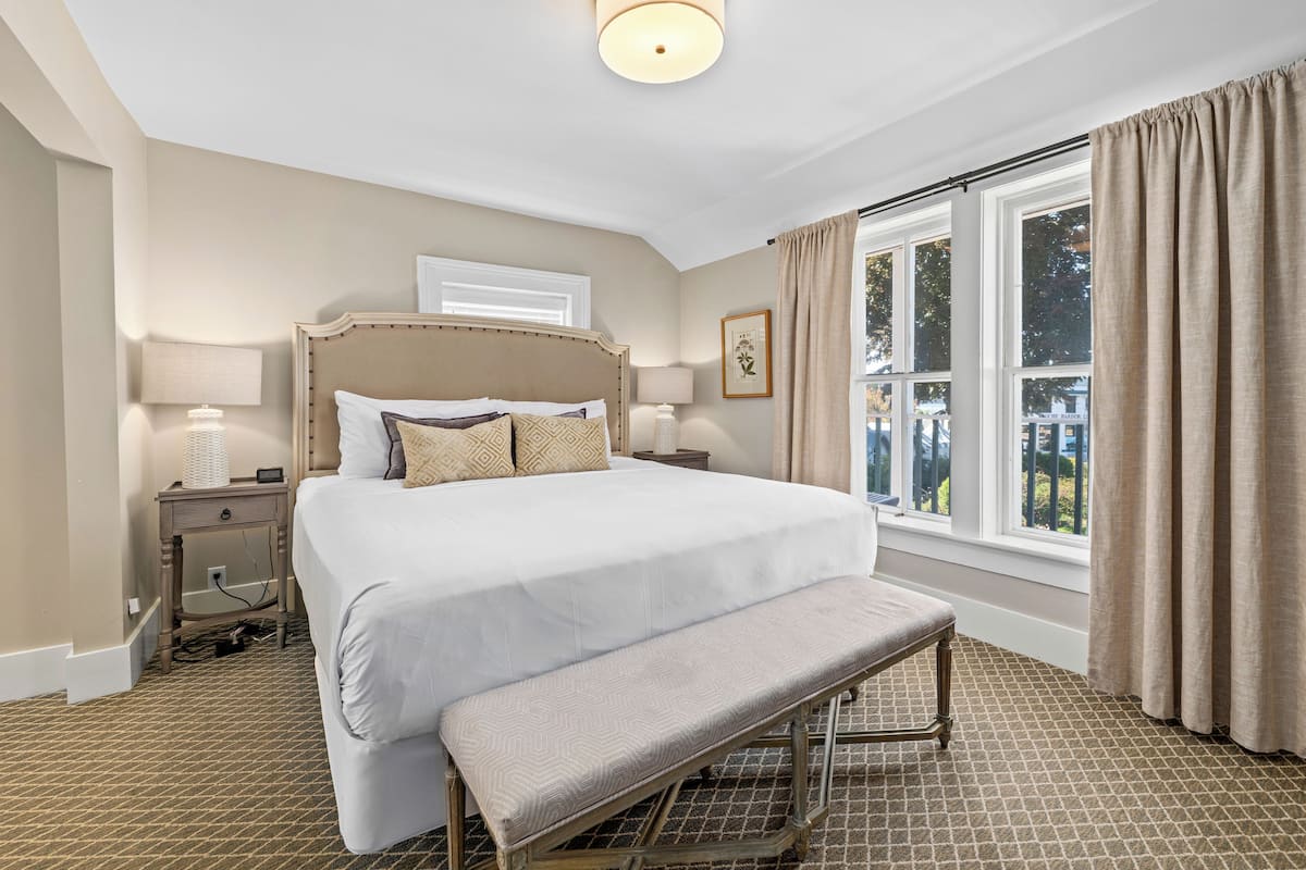 A neatly arranged bedroom with a large bed, a bedside table with a lamp, a bench, and a window with curtains.