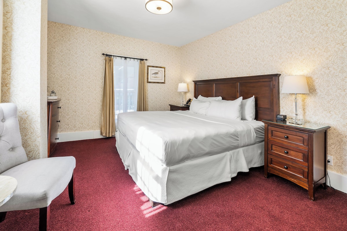 A hotel room with a large bed, a wooden nightstand with a lamp, a chair, and a window with curtains.