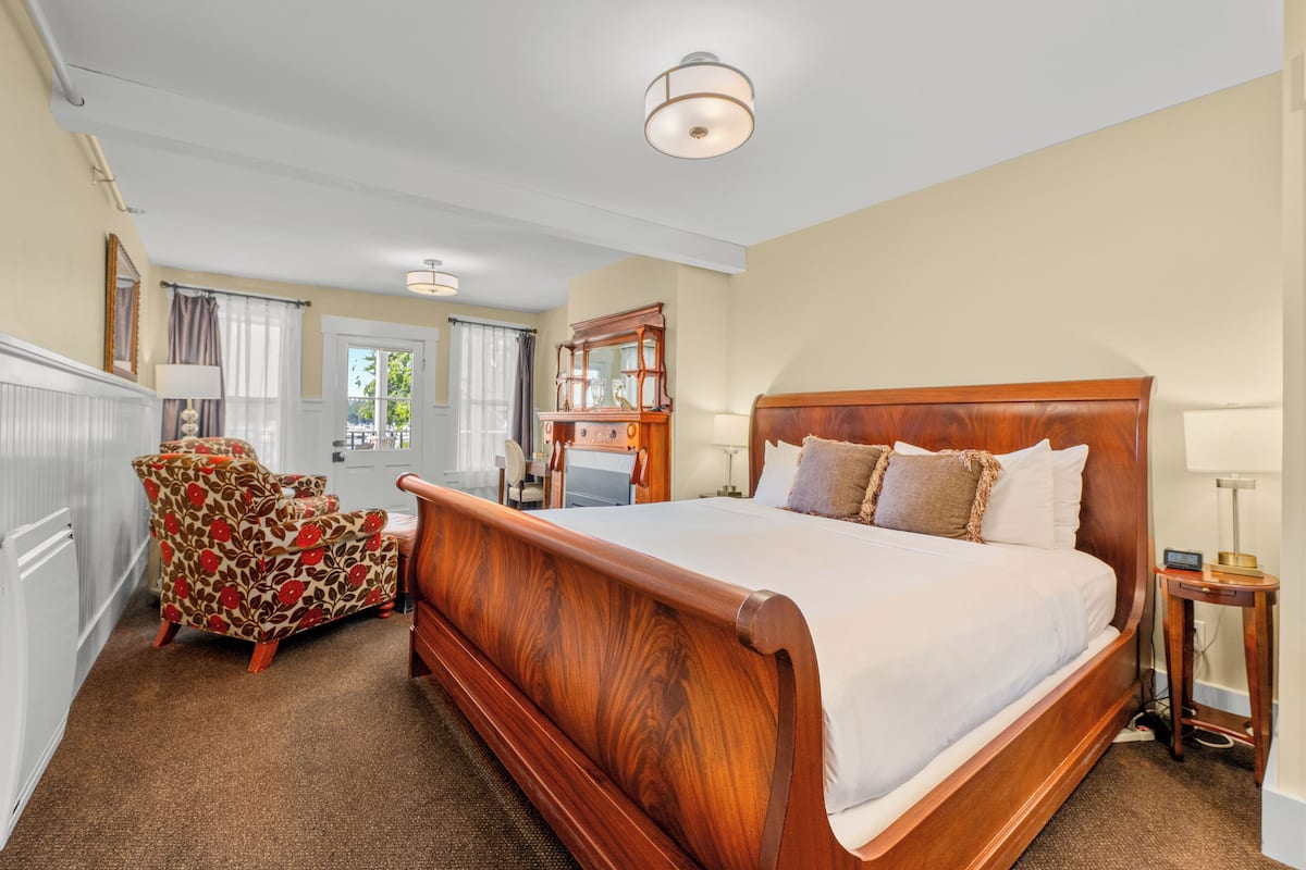 A bedroom with a wooden bed, a floral armchair, a wooden dresser, and a desk with a lamp.