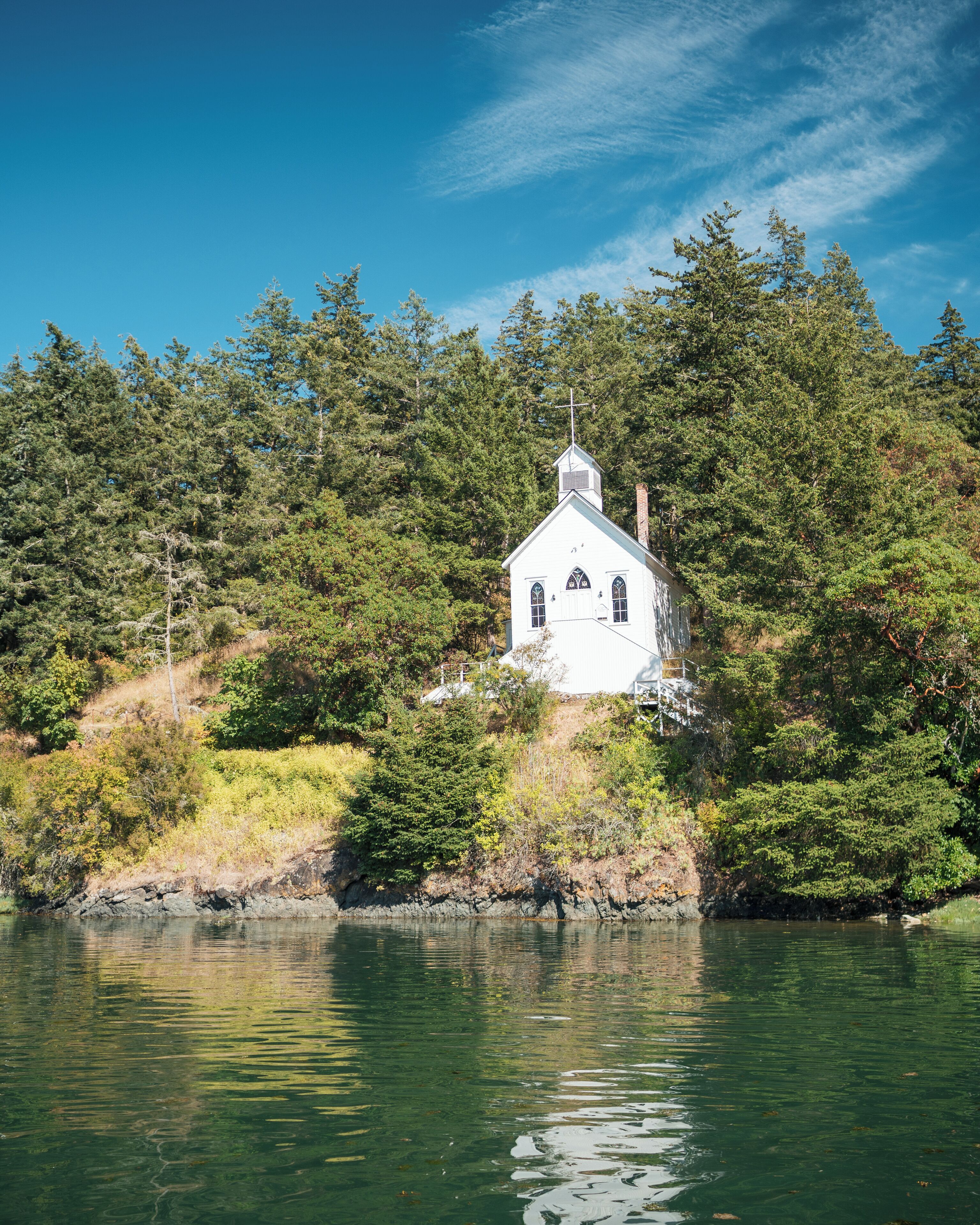 A white church with a cross on top, surrounded by dense forest and a body of water.