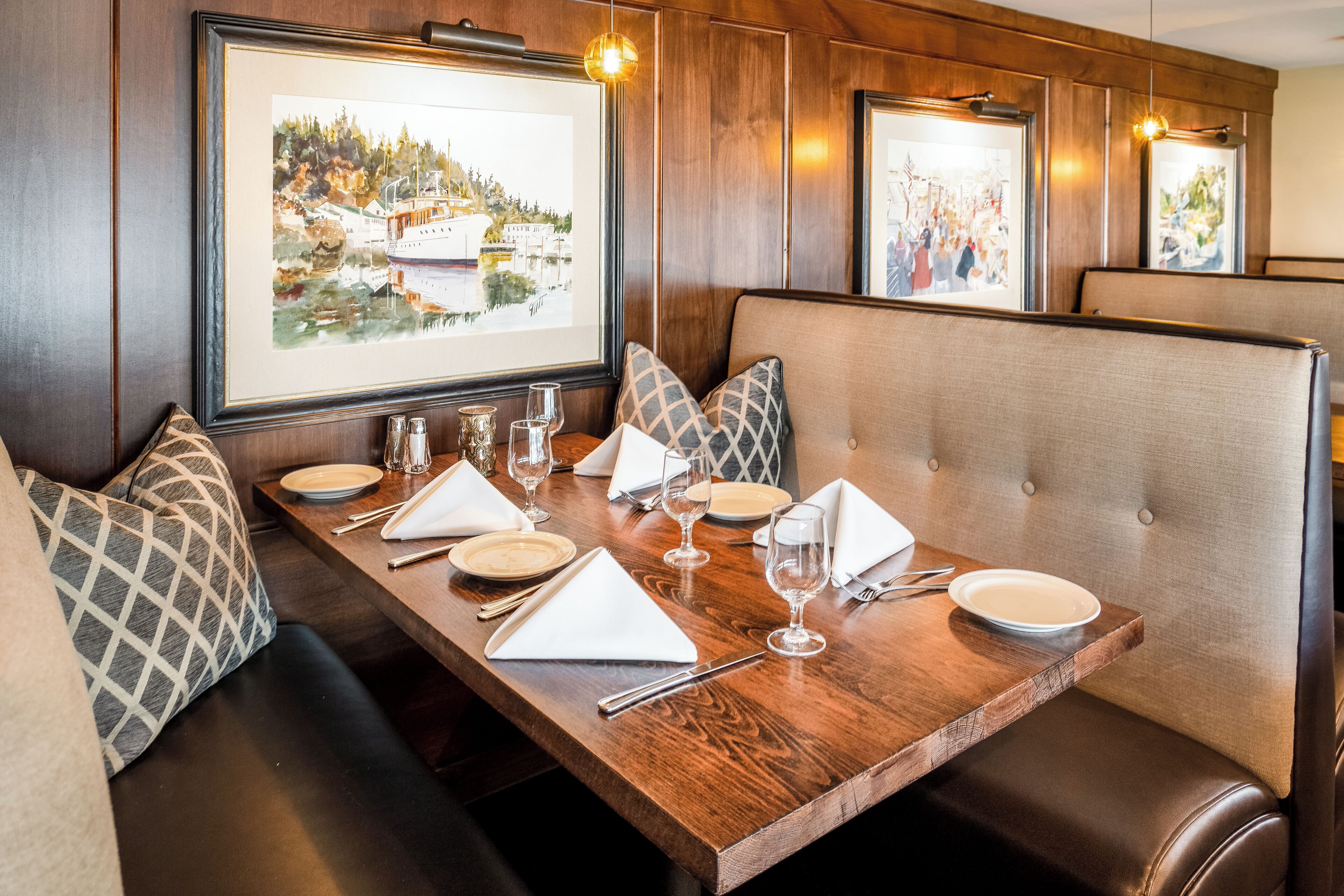 Uma área de refeições com mesa de madeira posta para quatro pessoas, assentos acolchoados e quadros decorativos nas paredes.