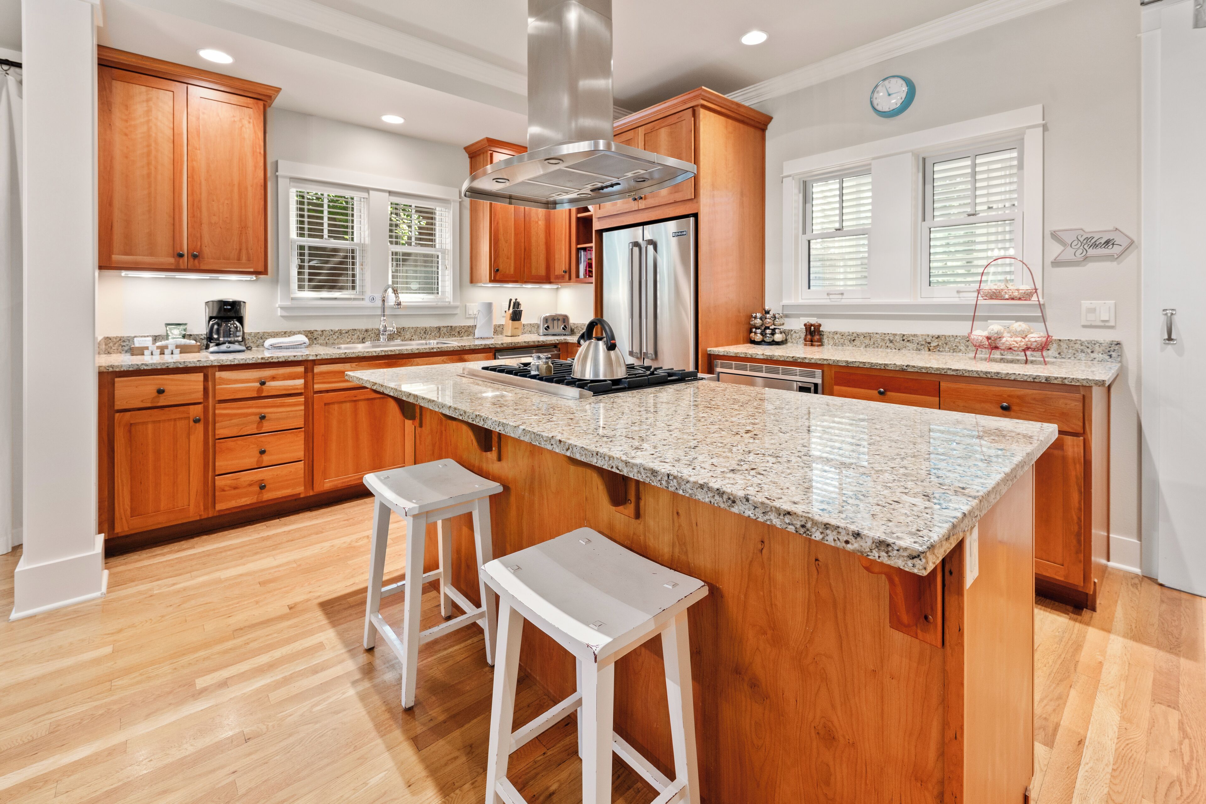 A kitchen with wooden cabinets, a stainless steel refrigerator, a granite countertop, and two white stools.
