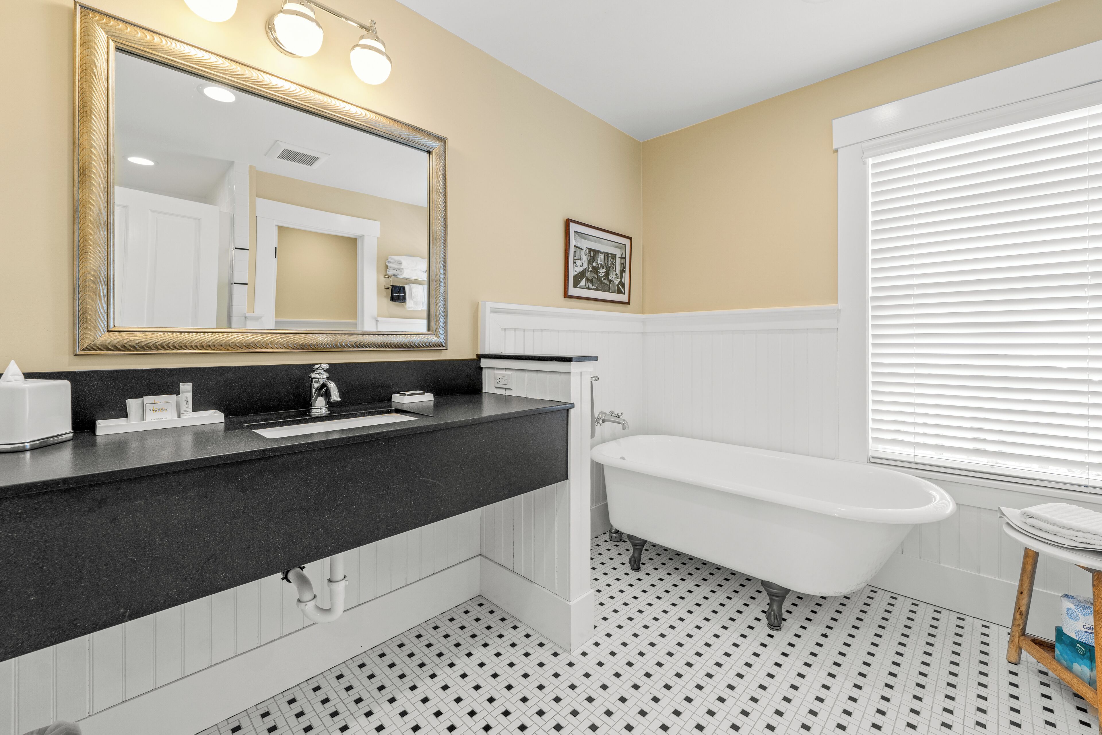 A bathroom with a black countertop, a white bathtub, a large mirror with lights, and a window with blinds.
