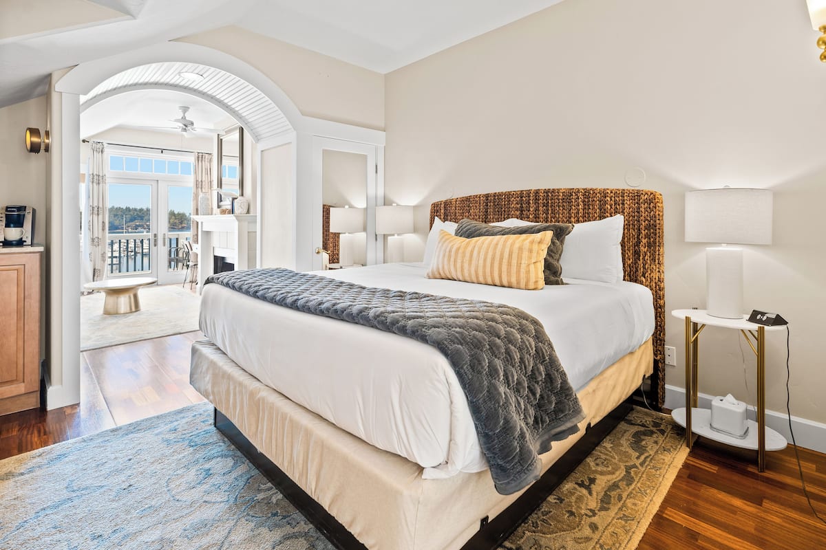 A spacious bedroom with a large bed, a bedside table, a nightstand, and a view of the outdoors.