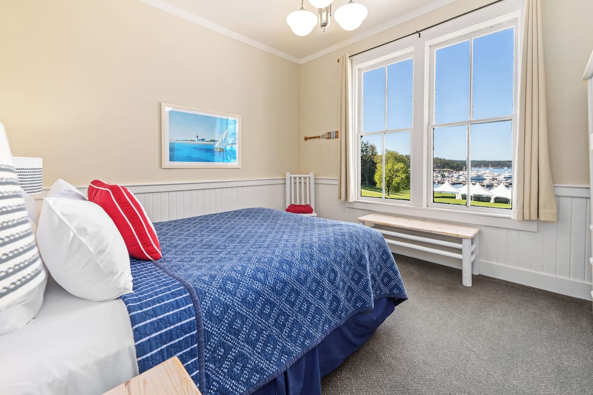 A bedroom with a large window, a bed with blue bedding, a red pillow, and a framed picture on the wall.