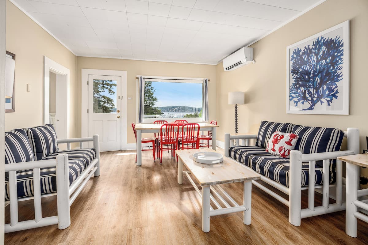 A room with a wooden floor, a dining table with chairs, a sofa, and a large window with a view.