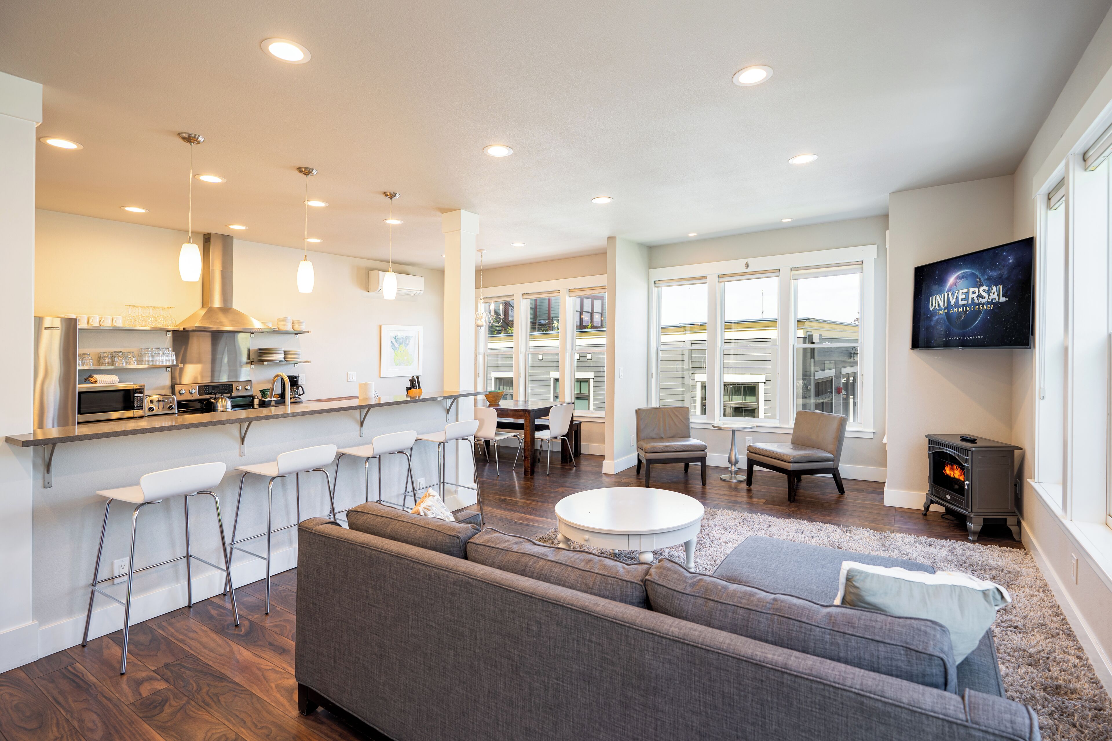 A modern kitchen with bar stools, a dining area with a round table, a living room with a sofa and a TV, and a fireplace.
