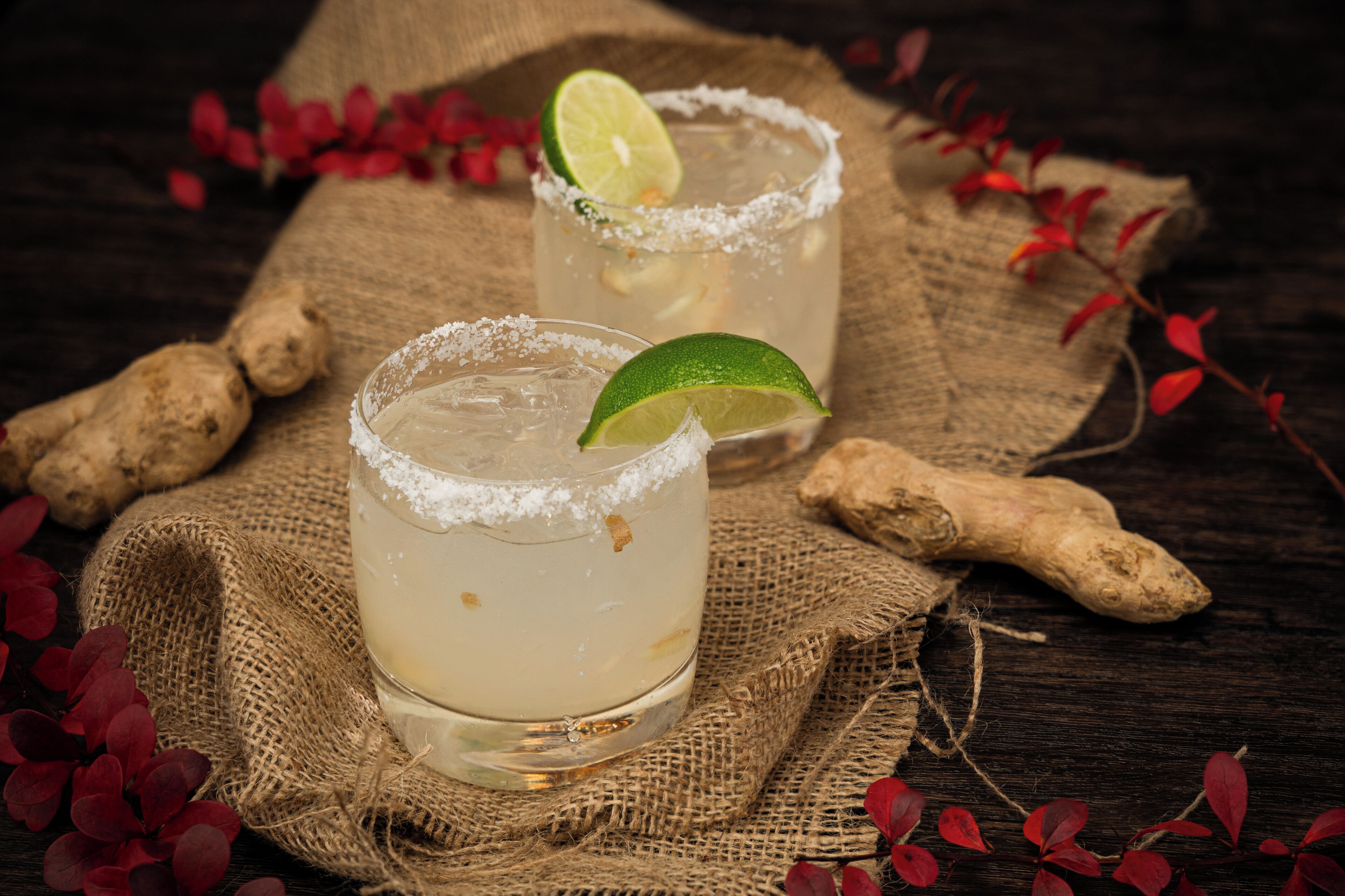 Two glasses of a clear beverage with lime garnish, placed on a burlap cloth with ginger roots and red leaves around them.