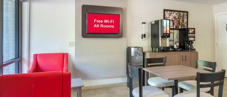 Habitación de hotel con comedor, una silla roja, cafetera y letrero que indica Wi-Fi gratuito en todas las habitaciones.